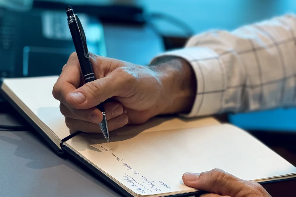Mãos anotando em um caderno com caneta, sobre a mesa, com notebook ao fundo